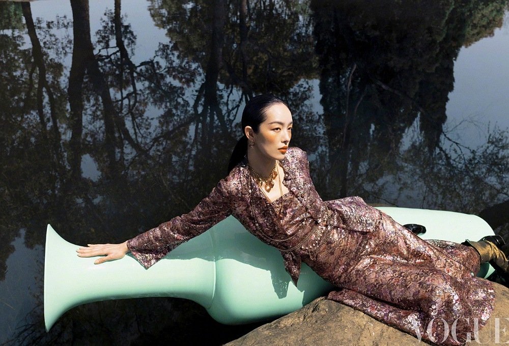 Woman in shimmering dress reclines on a large, light-green sculpture, posing in a Vogue fashion editorial with a woodland reflection.