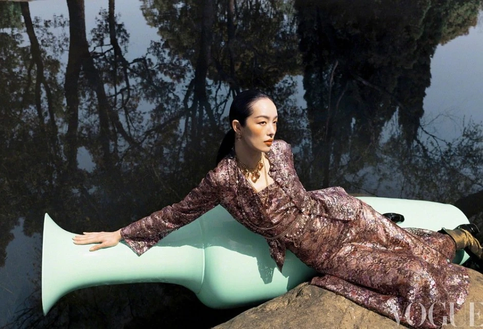 Woman in shimmering dress reclines on a large, light-green sculpture, posing in a Vogue fashion editorial with a woodland reflection.