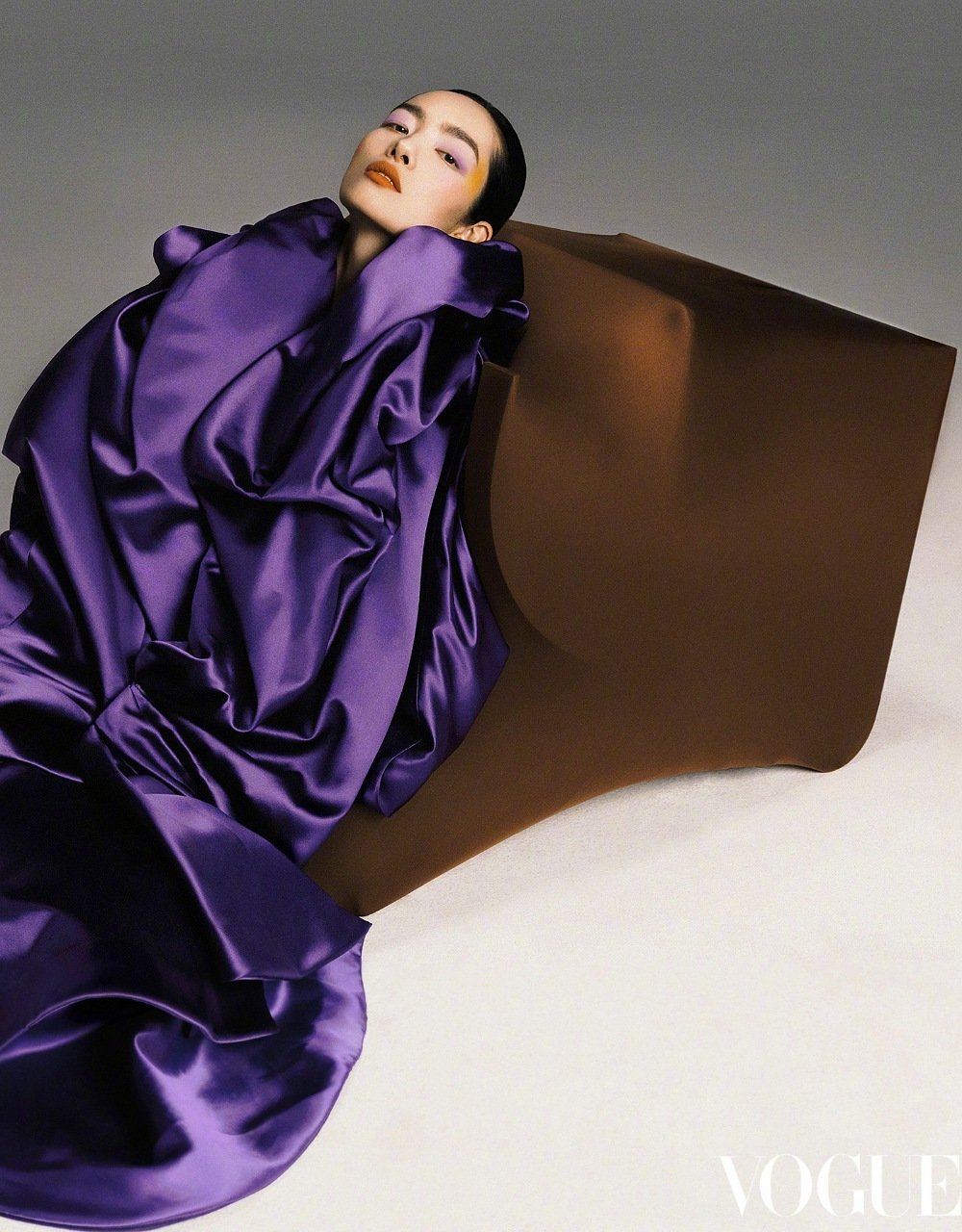 Fashion editorial shot of an Asian female model in a draped purple gown posing with a brown geometric chair against a grey backdrop.