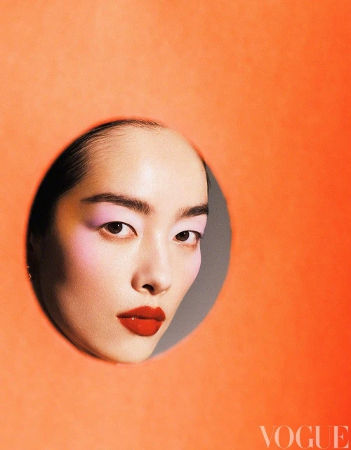 Headshot of an Asian female with pink eyeshadow and red lipstick, peering through a round orange paper cutout for beauty advertising.