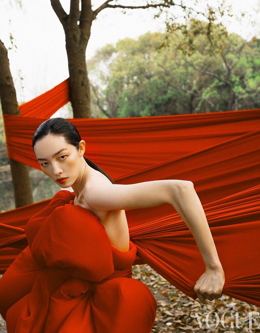 Model FeiFei Sun crouching in a sculptural red gown against draped crimson textiles wrapped around trees in a woodland setting.