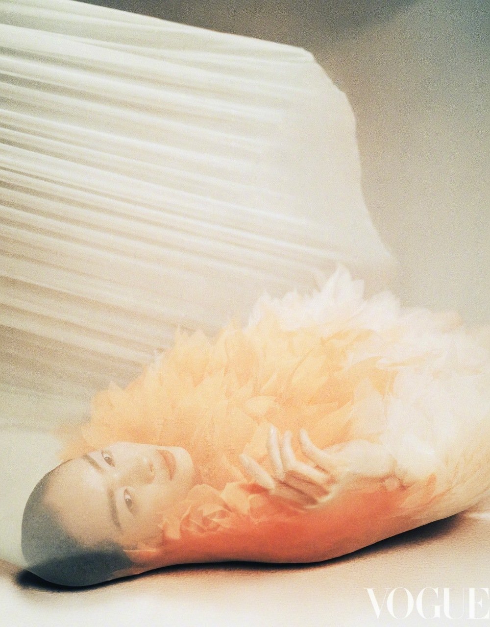 Woman with bald head lies in a feathered orange dress in front of a white pleated screen in a Vogue magazine beauty editorial.