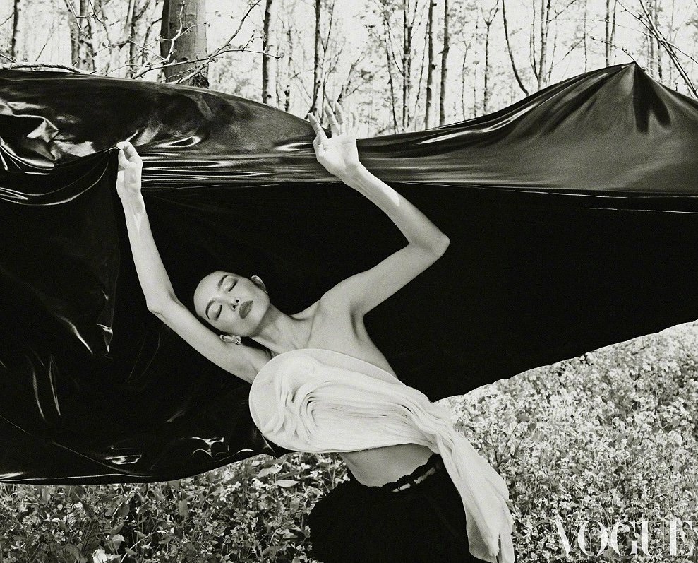 Monochrome fashion shot of a model lying in a meadow, draped in flowing black fabric and a white garment, near trees.