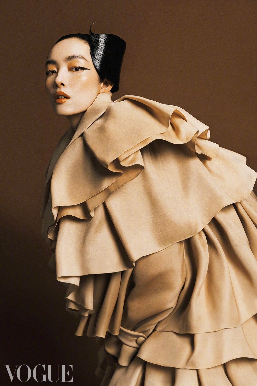 Editorial fashion shot of a stylish Asian woman in beige tiered ruffle coat and glossy black hairpiece against a neutral brown background.