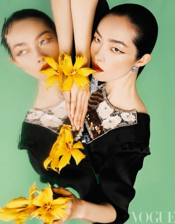 A woman with slicked-back hair and dark eye makeup poses with a yellow flower against a green background, reflected in a glossy surface.