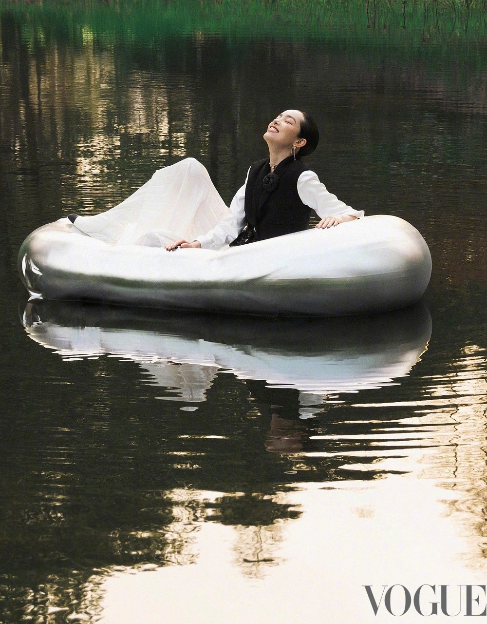 Woman in white dress and black vest enjoys a boat ride on a tranquil lake with reflections, captured in soft, natural light.