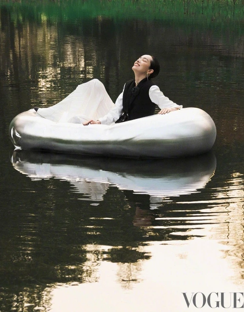 Woman in white dress and black vest enjoys a boat ride on a tranquil lake with reflections, captured in soft, natural light.