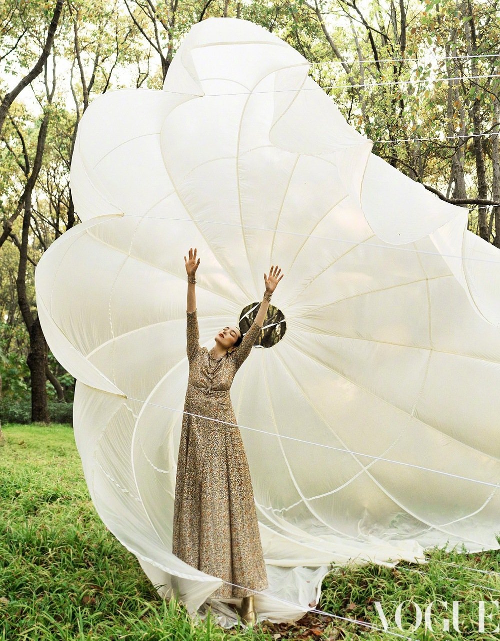 Woman in patterned maxi dress poses under white parachute in grassy outdoor setting with forest backdrop. Vogue logo in corner.