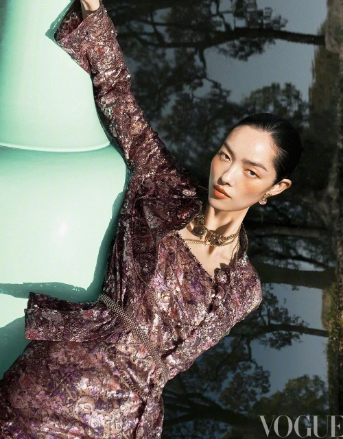 Fashion editorial photo of an adult woman in a shiny purple dress and gold jewelry, posing outdoors near trees and green abstract object.
