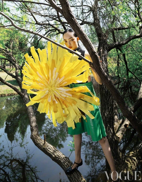 Woman in green skirt stands on tree branch near water, holding a large yellow flower, with a background of trees and vegetation.