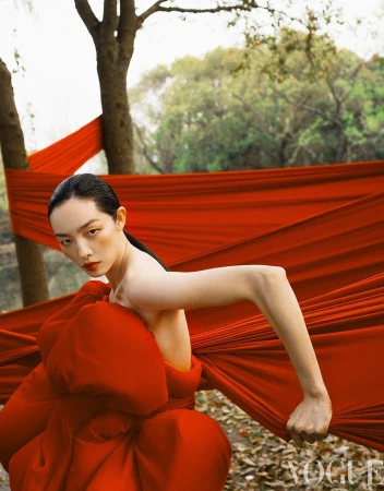 Model FeiFei Sun crouching in a sculptural red gown against draped crimson textiles wrapped around trees in a woodland setting.