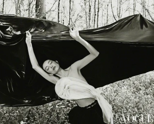 Model FeiFei Sun holding a glossy black fabric sheet outdoors in a wooded setting for Vogue China editorial photography.