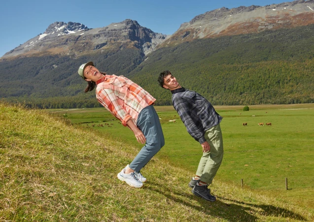 Two people in plaid shirts laugh on a grassy hillside with mountains and green fields in the background on a sunny day in New Zealand.