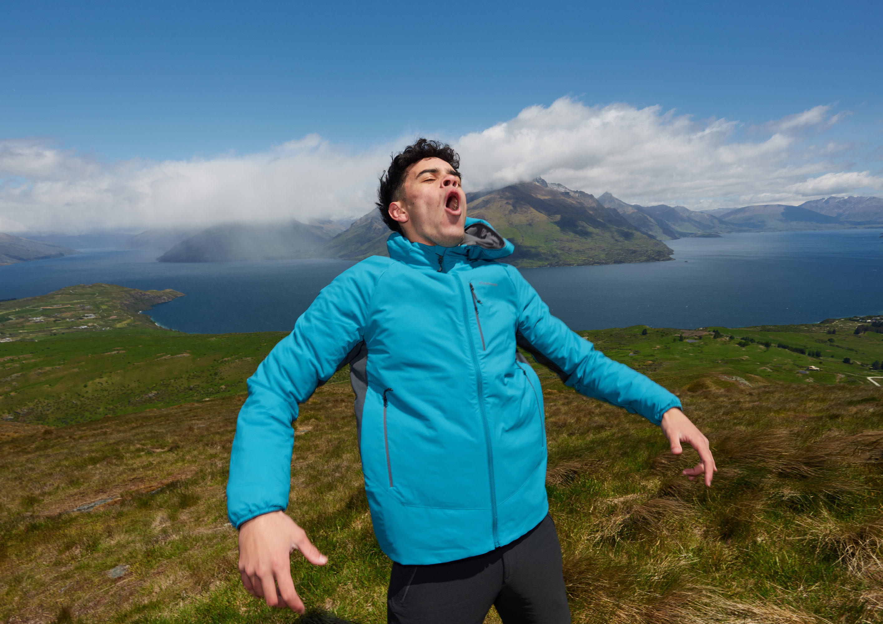 Person in blue puffer jacket with open mouth in grassy mountain landscape with lake and mountains under a partly cloudy blue sky.