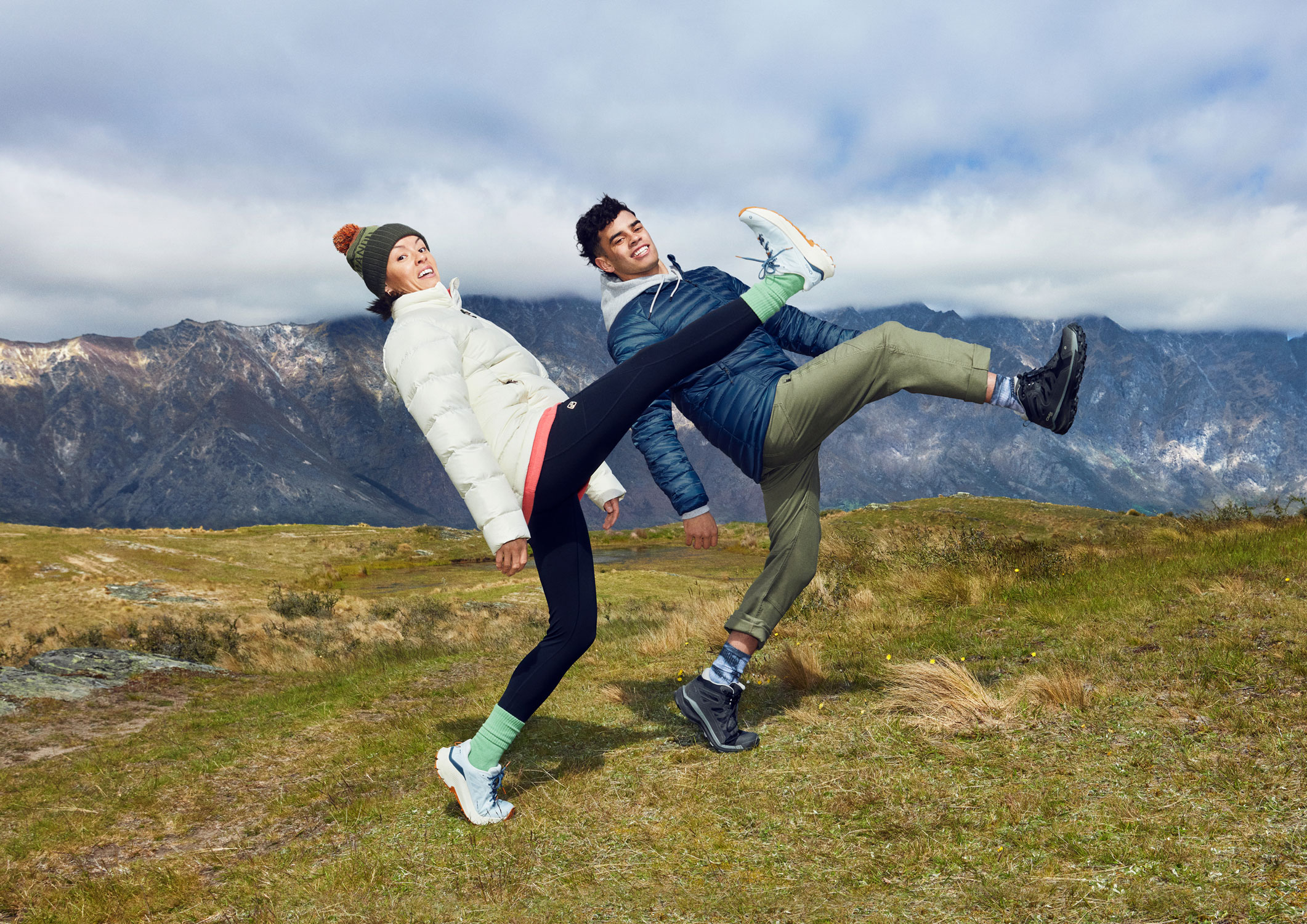 Two people kicking in athletic wear in a grassy field with mountains in the background, wearing jackets, hats, leggings, and hiking shoes.