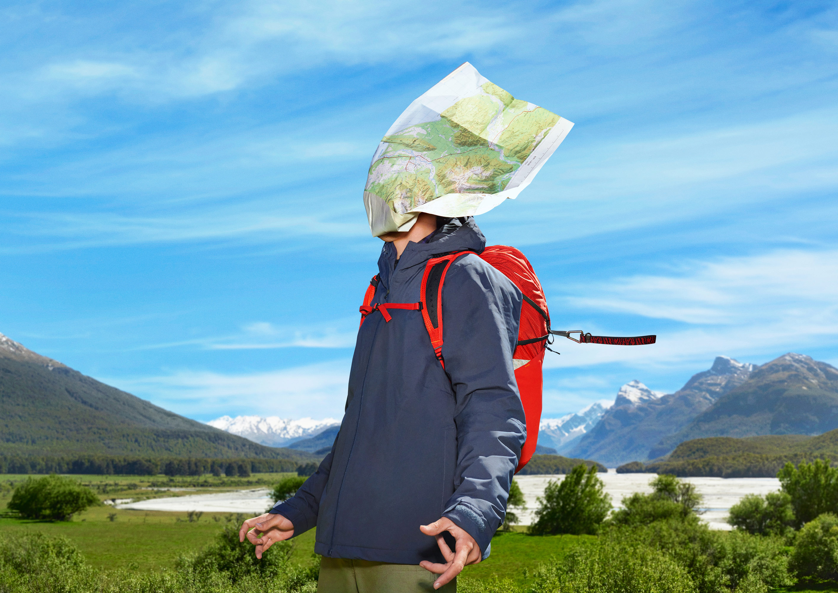 Person with a map over their head wearing a blue jacket and red backpack stands in a mountain landscape on a sunny day.