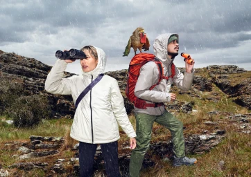 Two hikers in waterproof jackets exploring a rocky landscape while a kea bird carrying a red wallet perches on a backpack