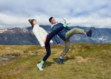 Two people outdoors in outdoor apparel kicking legs up playfully with a mountainous landscape behind them under a cloudy sky.