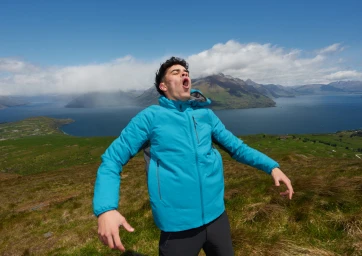 Man in a bright blue jacket yelling with arms outstretched on a grassy hill overlooking a lake and mountains