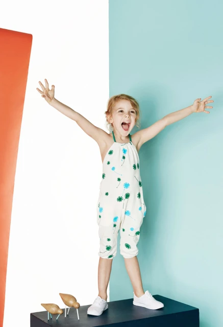 A blonde child in a romper stands atop a dark blue platform with open arms in a colorful corner setting with modern wood bird figures.
