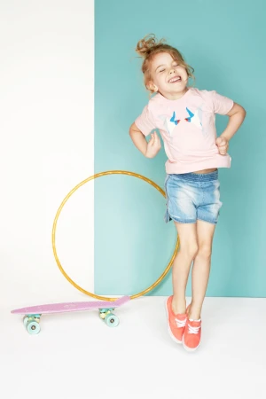Excited young girl jumping mid-air in a studio with white and blue backdrop, featuring a skateboard and hula hoop. 