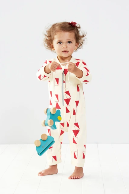 Adorable baby girl in patterned jumpsuit stands indoors holding a wooden toy, captured in a bright, full shot on a white background.