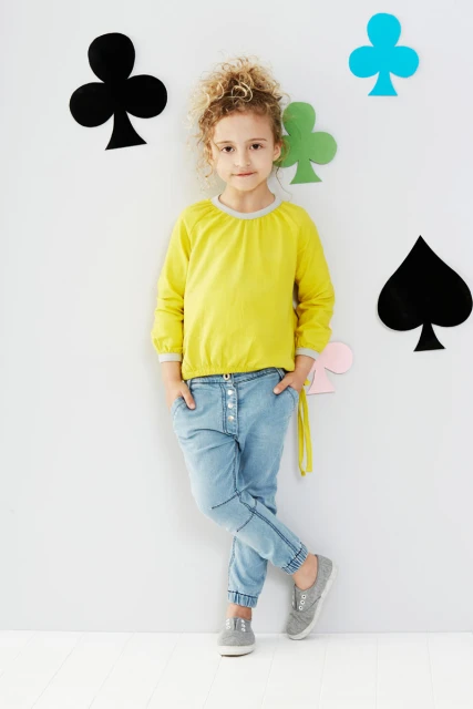 A young girl with curly blonde hair poses in a yellow long-sleeve shirt and blue jeans in front of a white wall with card suit shapes.