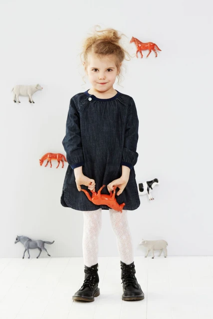 Adorable little girl stands in a denim dress, holding a red animal toy, with toy animals affixed to the white wall behind her.