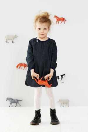 Adorable little girl stands in a denim dress, holding a red animal toy, with toy animals affixed to the white wall behind her.