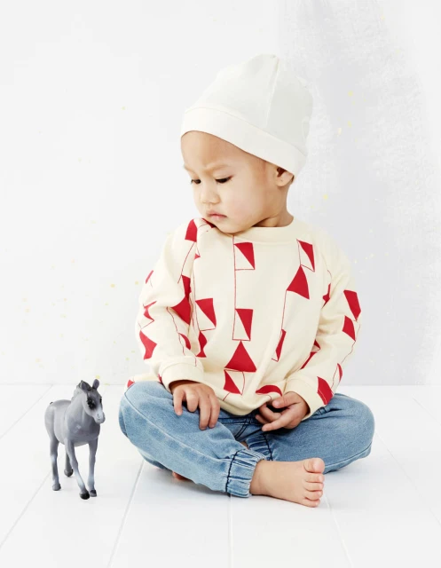 Toddler wearing a beanie sits on a white floor next to a gray toy horse, looking down with a neutral expression indoors.