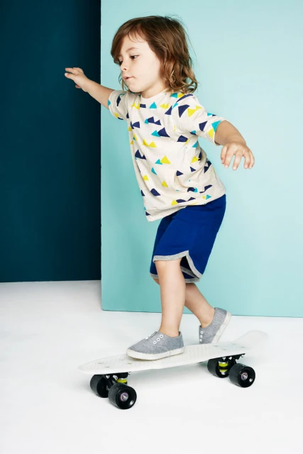 A young child with long hair is riding a white skateboard indoors in a brightly lit studio with a blue backdrop and a white floor.