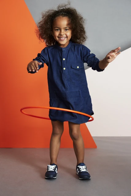 Smiling child with curly hair playing with an orange hula hoop in a studio with an orange and gray background.