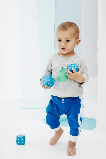 A toddler wearing blue pants and a grey shirt holds blue polka-dotted dice in a white photo studio with translucent blue panels.