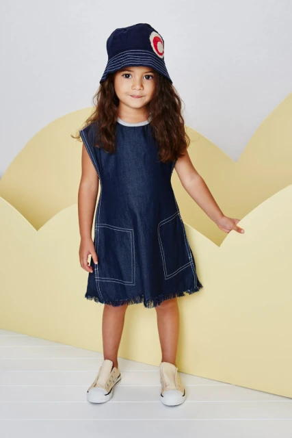 Little girl in a blue denim dress and matching hat poses in a studio with a yellow, cloud-shaped prop and a white background.