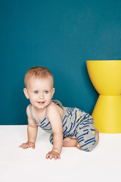 Baby on all fours in gray patterned jumpsuit against teal wall with yellow stool. Playful studio shot with focus on baby.