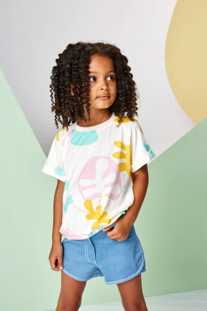 Cute little girl with curly hair in a patterned T-shirt and denim shorts posing indoors against a split-tone pastel background.