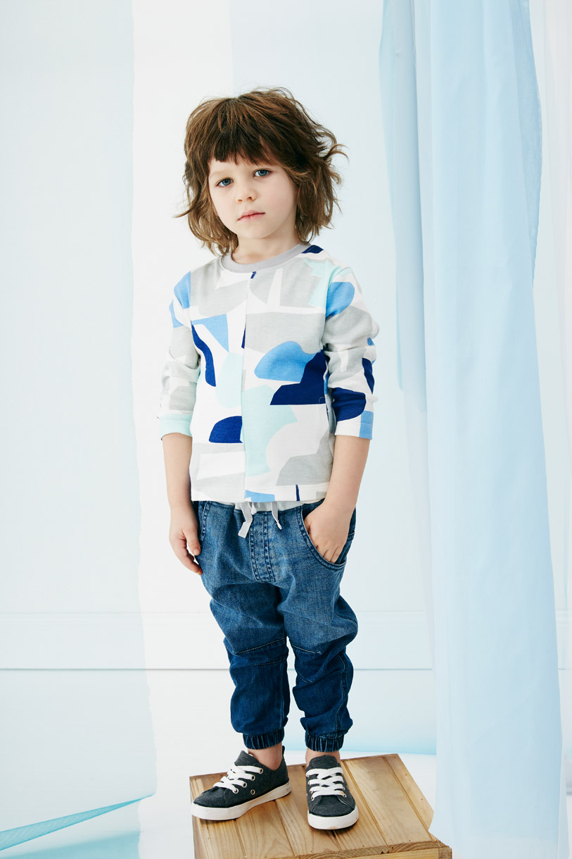 A young child with long brown hair stands on a wooden platform wearing a long-sleeved shirt, denim pants, and black sneakers in a studio.