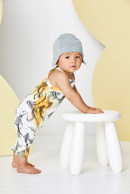 Adorable toddler in patterned romper and grey beanie leans on white stool in a cheerful studio setting with soft yellow background.