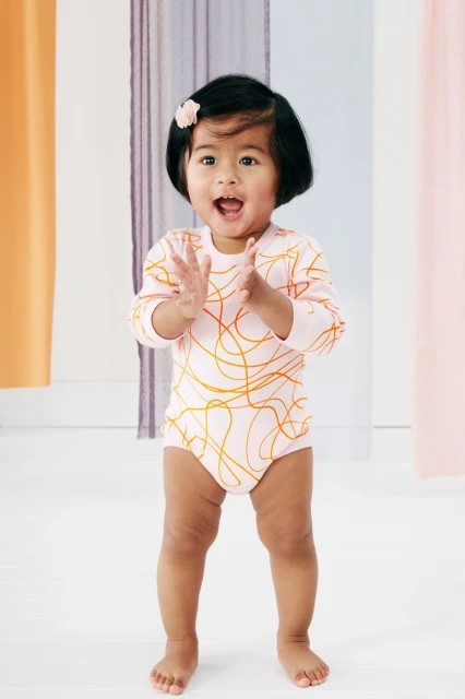 Full-length shot of a happy toddler clapping her hands, wearing a patterned bodysuit in a light-filled indoor setting with colorful curtains.