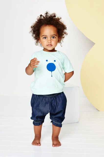 Toddler in a light green shirt with a blue bear face graphic standing barefoot in front of a white and yellow background.