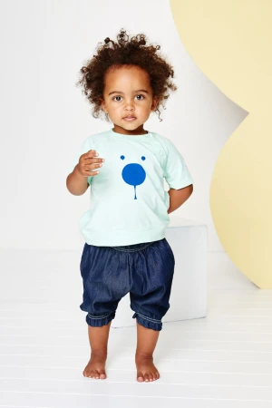 Toddler in a light green shirt with a blue bear face graphic standing barefoot in front of a white and yellow background.