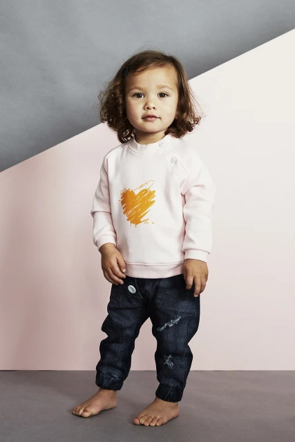 Full-length portrait of a toddler with curly brown hair, wearing a pink sweatshirt with an orange heart and dark denim jeans.