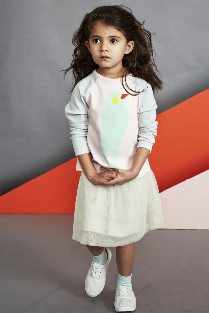 Full shot of a child in a tutu skirt and printed shirt, facing forward, on a patterned gray and orange studio background.