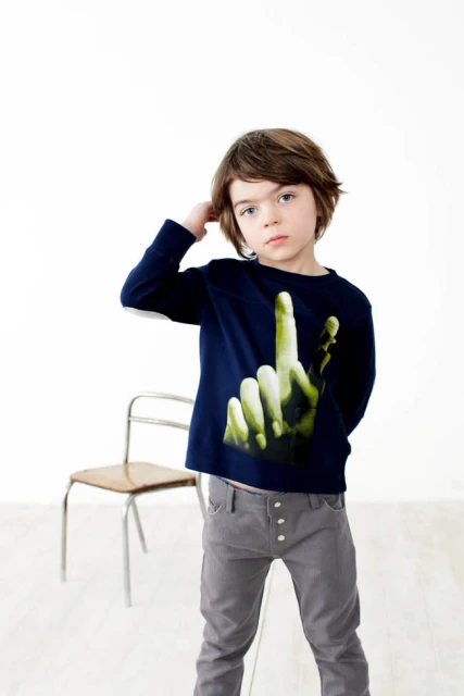 Full shot of a child standing in a studio. The child has brunette hair and is wearing a long-sleeved shirt with a graphic hand.