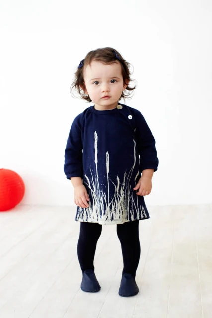 Full shot of a toddler wearing a dark blue dress with white grass-like pattern on a white background with a red ball on the side.