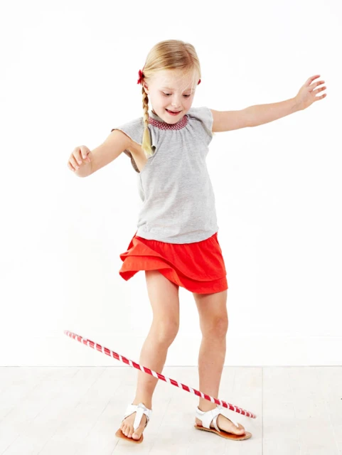 Young girl with blonde hair spins a hula hoop in a bright, white studio setting. She wears a grey top and red skirt.
