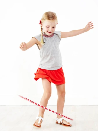Young girl with blonde hair spins a hula hoop in a bright, white studio setting. She wears a grey top and red skirt.