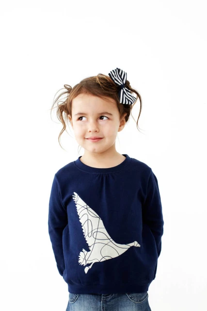 Adorable young girl with pigtails wearing a navy sweatshirt with bird design and jeans against a white background in a studio shot.