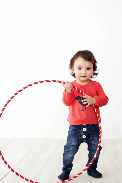 A young child plays with a red and white hula hoop, wearing a red shirt and denim pants in a brightly lit indoor setting.