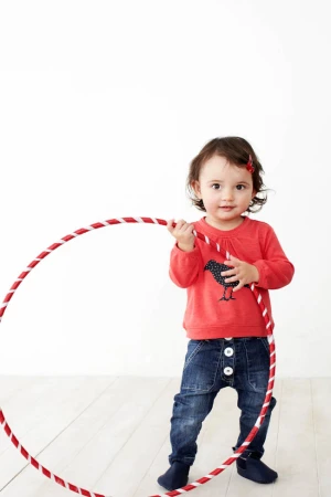 A young child plays with a red and white hula hoop, wearing a red shirt and denim pants in a brightly lit indoor setting.